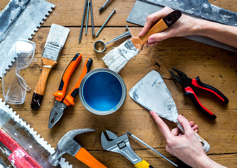 A wood table with painting supplies and tools with hands holding a paint brush and mud scraper