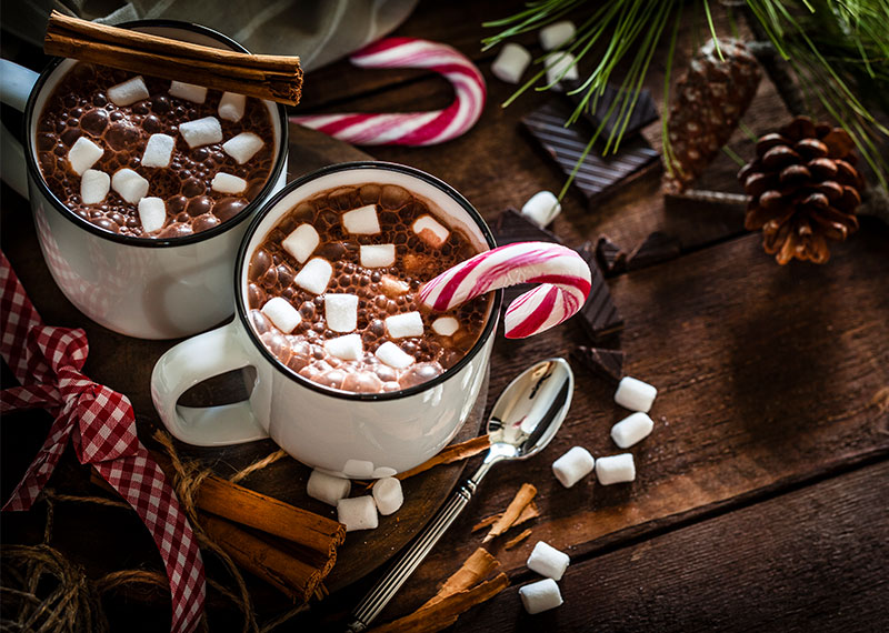 Two cups of hot cocoa with marshmallows and candy canes sitting on a wood table with pine cones around