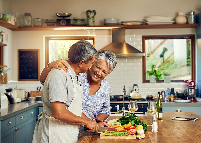 A husband and wife cutting vegetables with the wife's arm around the husband laughing