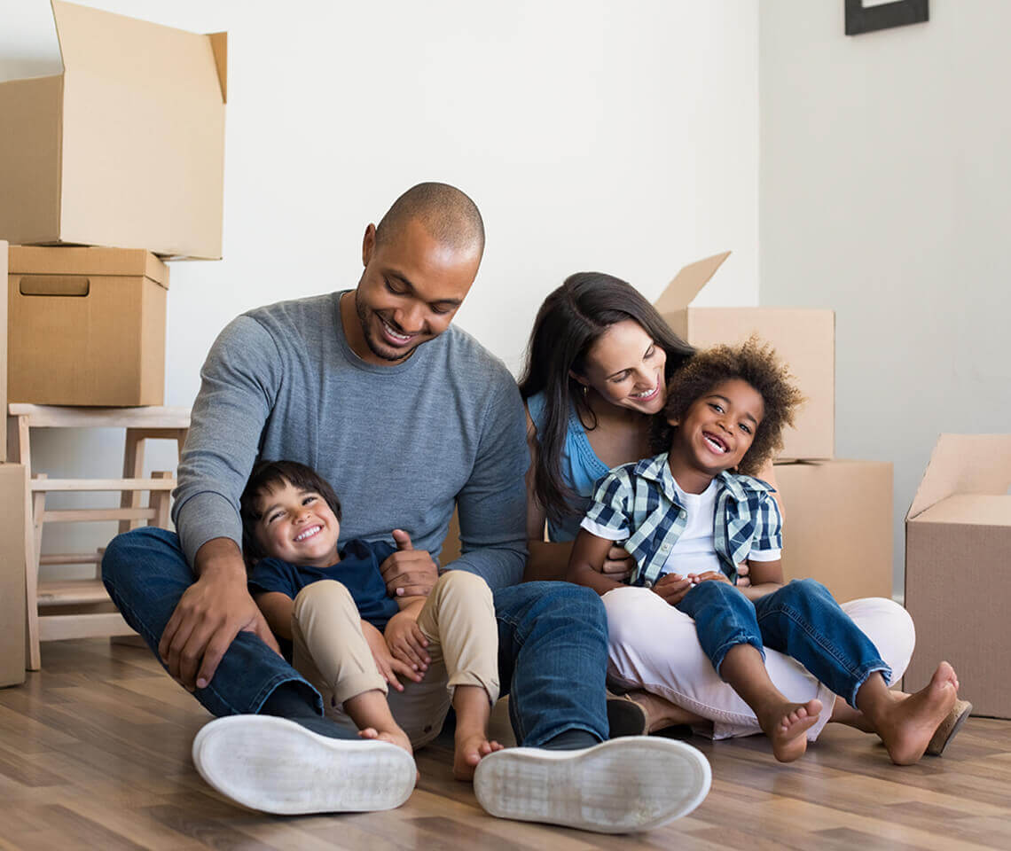 Family of 4 sitting on wood floor laughing with boxes surrounding them